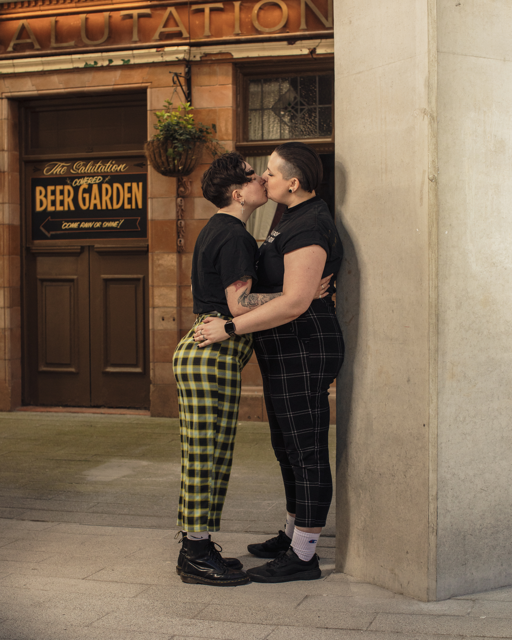 Two people kiss against a column on a street, a beer garden sign can be seen in the background.