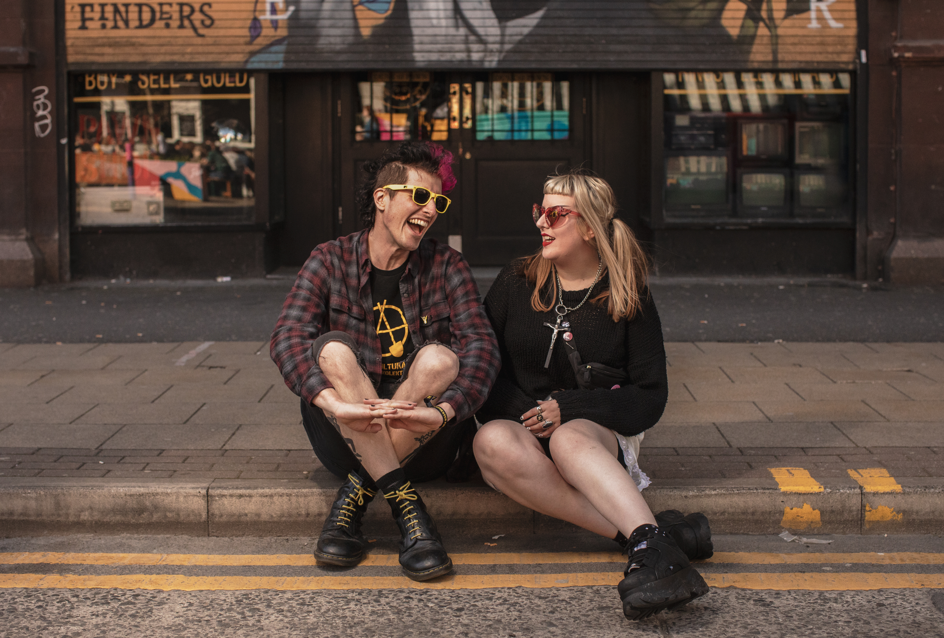 A couple sits on a sidewalk curb, the person on the left is laughing.