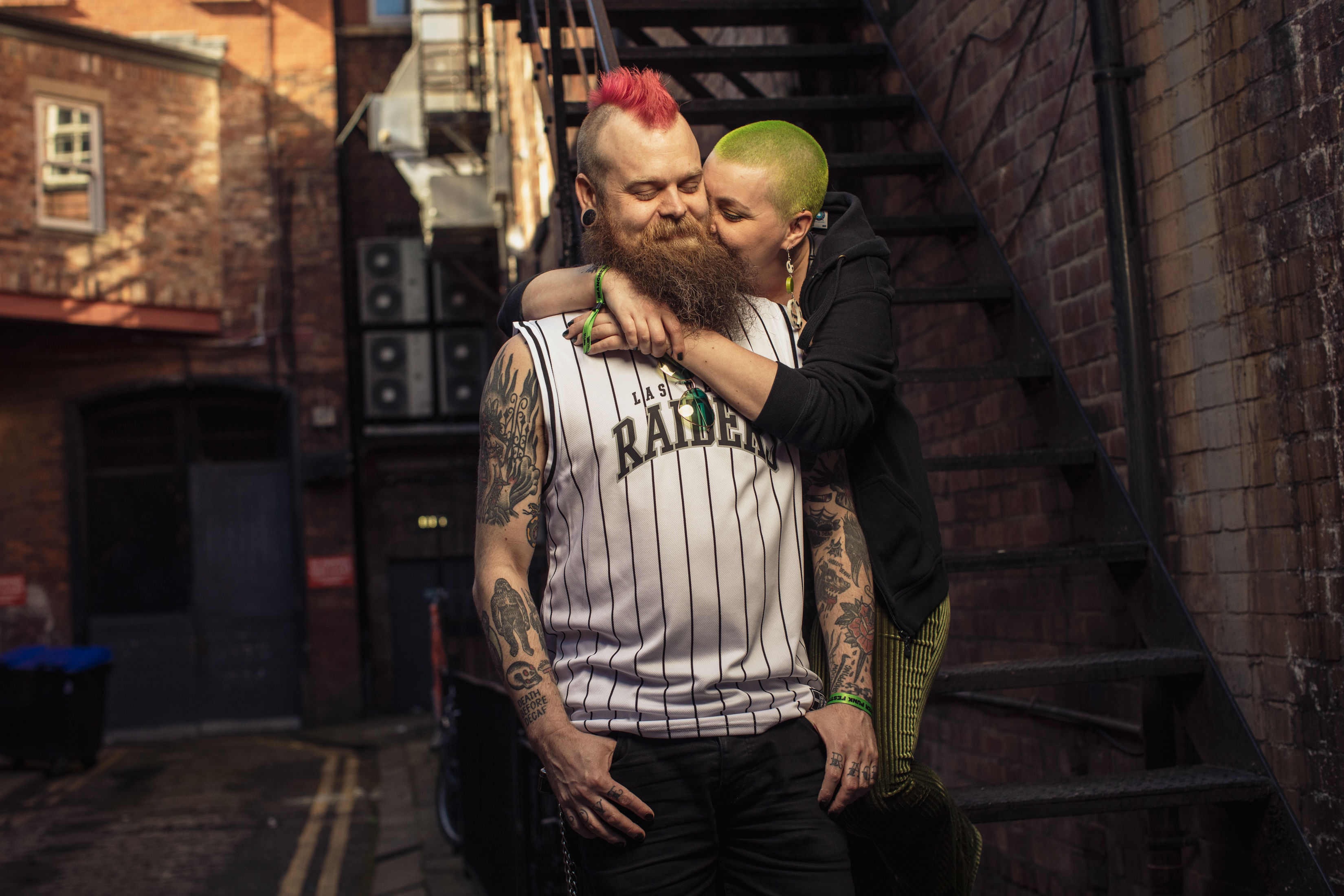 A man with a pink mohawk, beard, and an Raiders tank-top is being embraced from behind by a woman with a shaved, green hair.