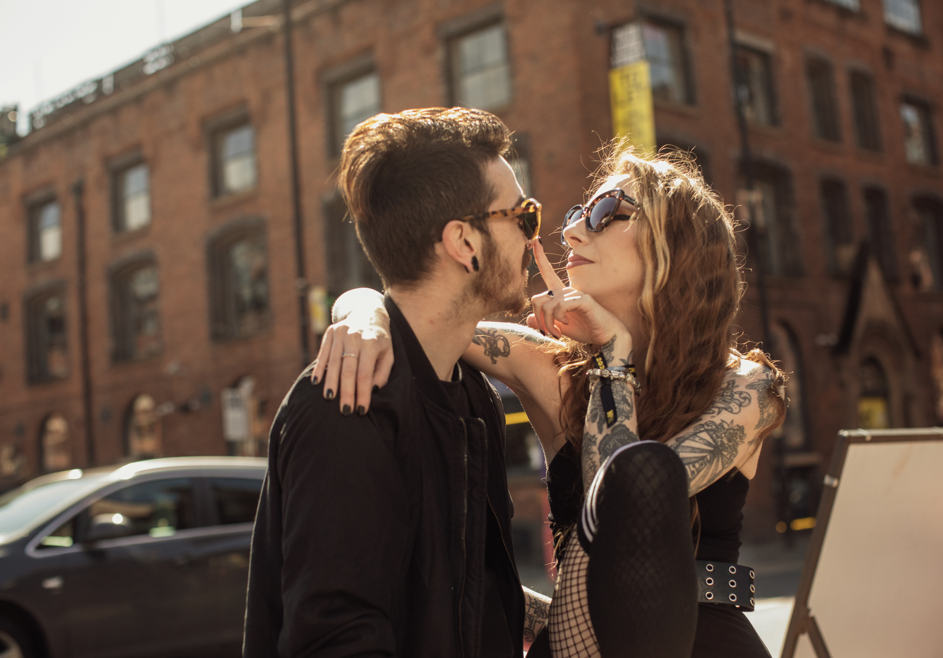 A couple looks into each others eyes through their sunglasses, the woman puts her finger on his nose in a flirty manner.